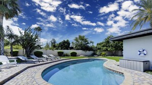 A swimming pool with a curved edge, surrounded by a brick patio, palm trees, and a white building with a life preserver on the wall.