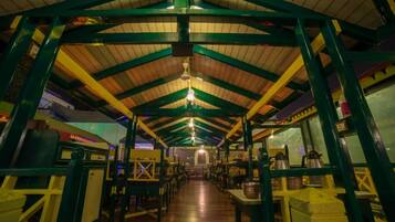 A restaurant with a wooden ceiling and green structural beams, featuring tables and chairs arranged for dining.