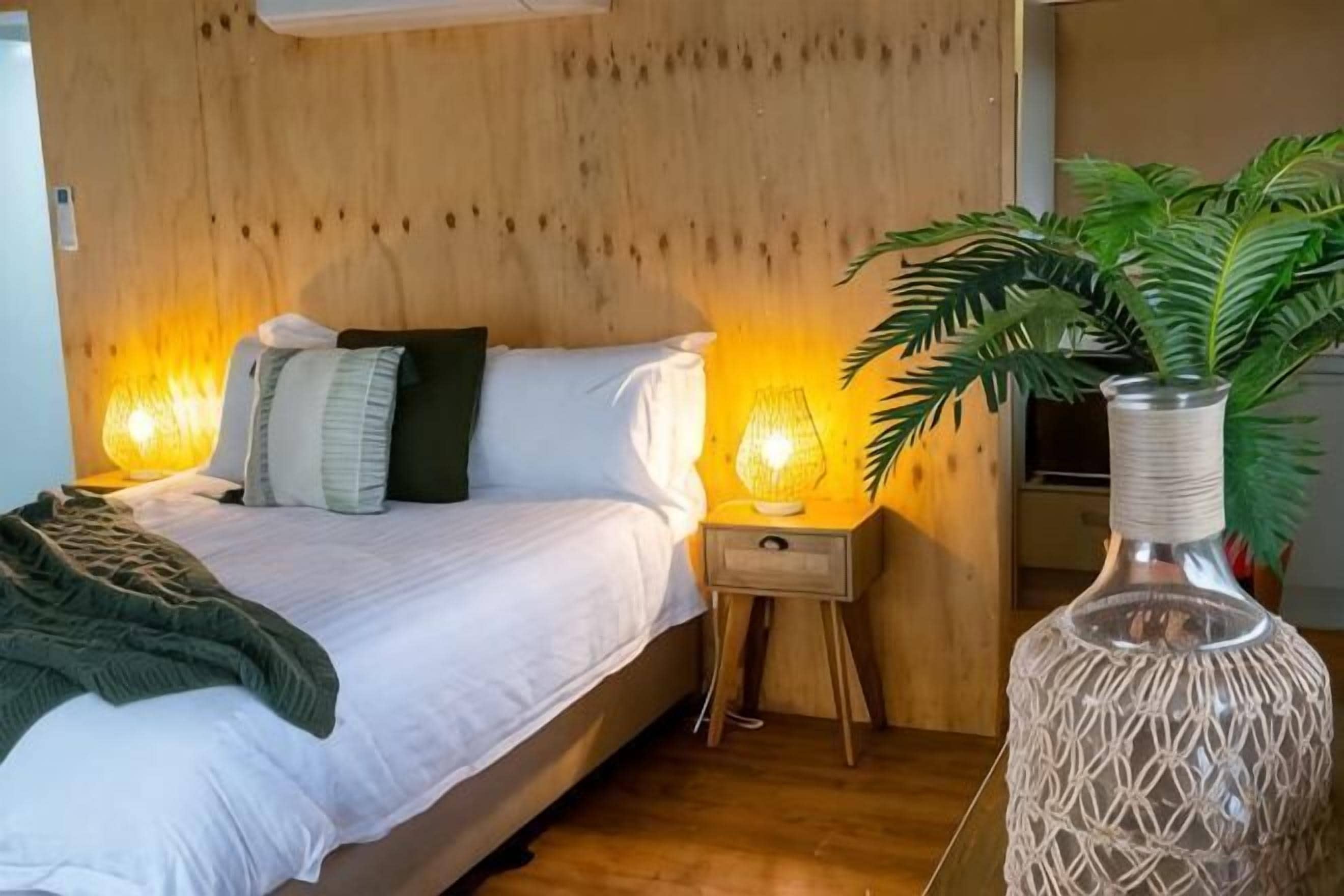 Un dormitorio con cabecera de madera, cama con ropa de cama blanca, dos lámparas de noche, una mesita de noche, un jarrón tejido y una planta en maceta.