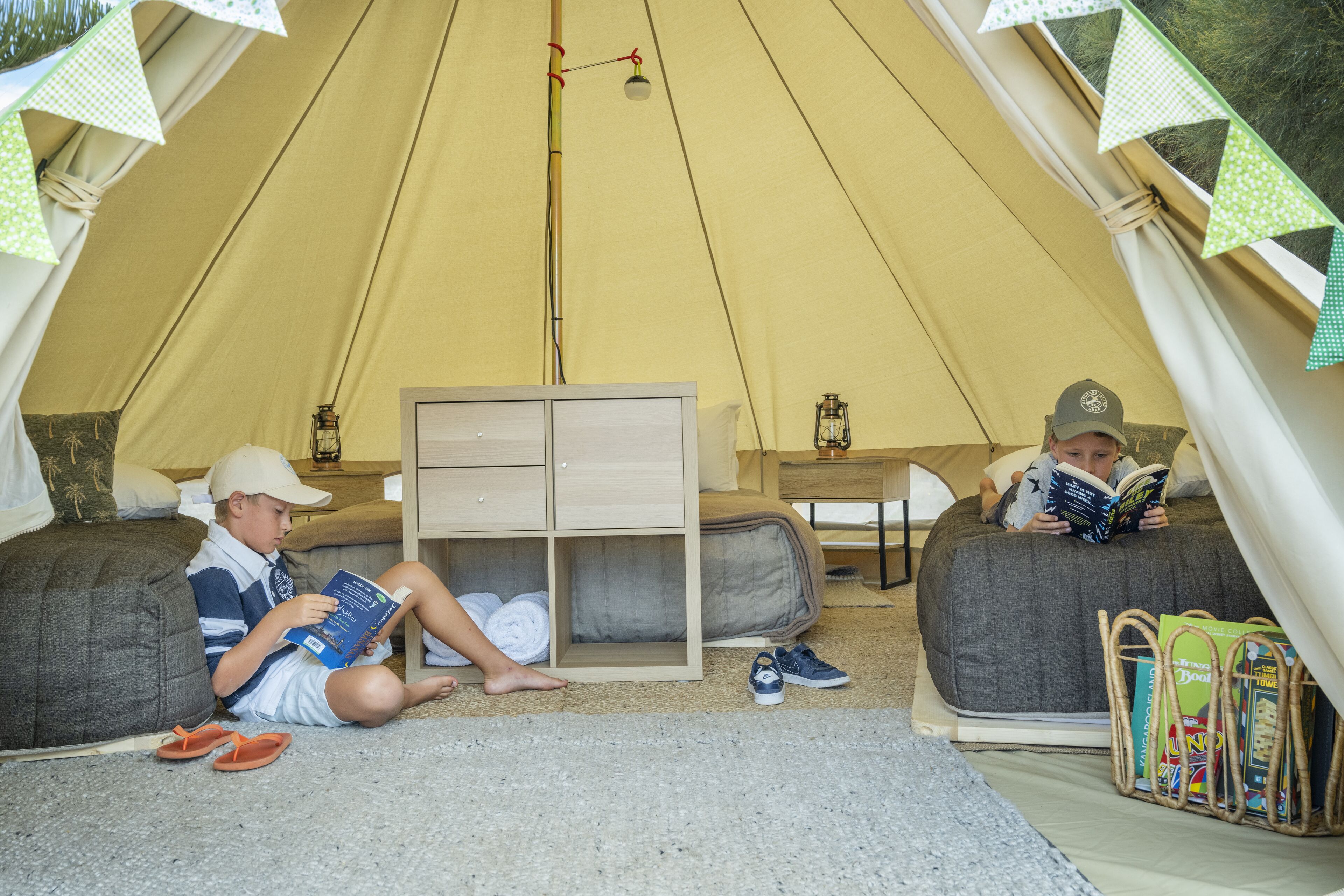 A tent with two sleeping areas, each with a pillow and a small table. There is a shelf with books and a lantern.