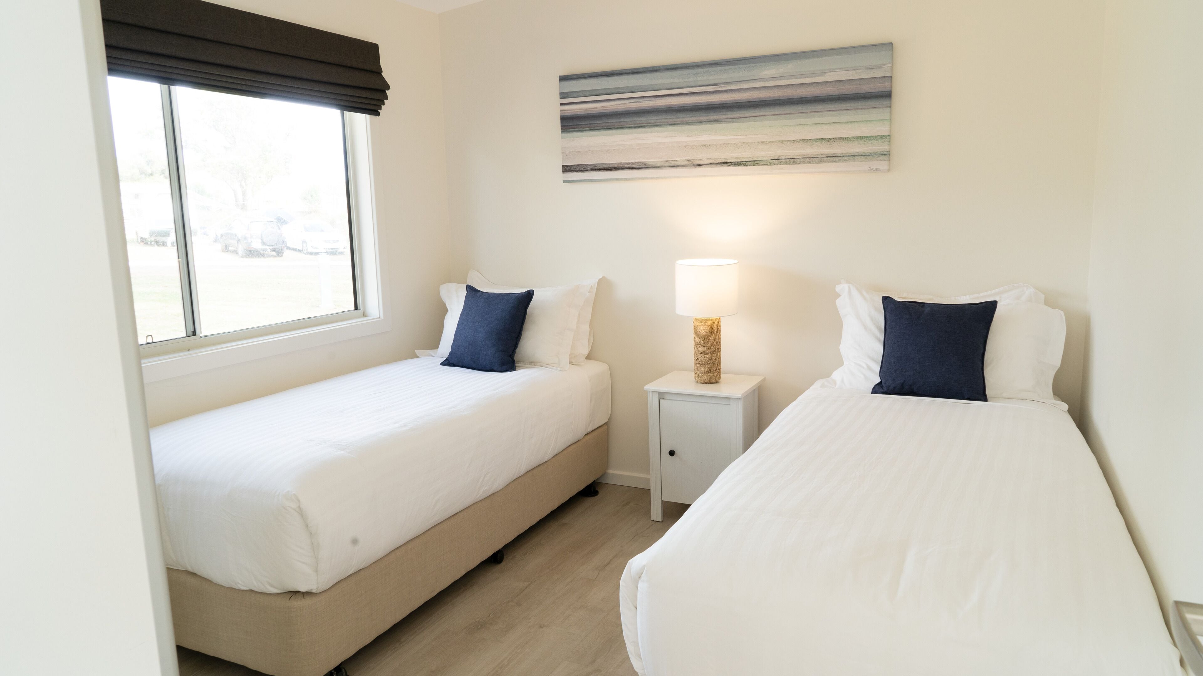 A hotel room with two single beds, a nightstand with a lamp, and a painting on the wall.