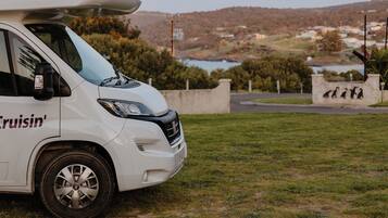Una caravana blanca con la frase 'Cruisin'' en el lateral estacionada en una zona cubierta de césped, con vistas panorámicas a colinas y una masa de agua al fondo.