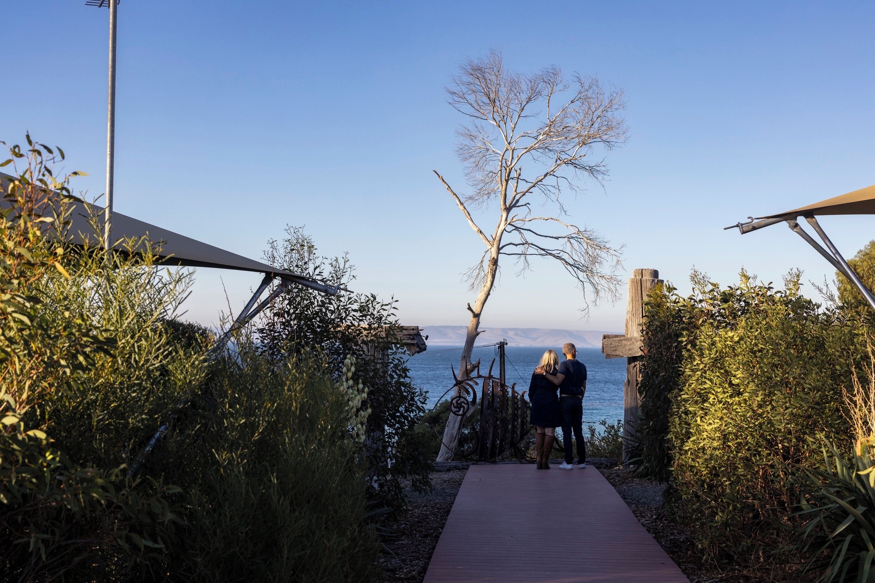 A pathway leading to a beach with a clear sky, a wooden fence, and a couple standing by the water.
