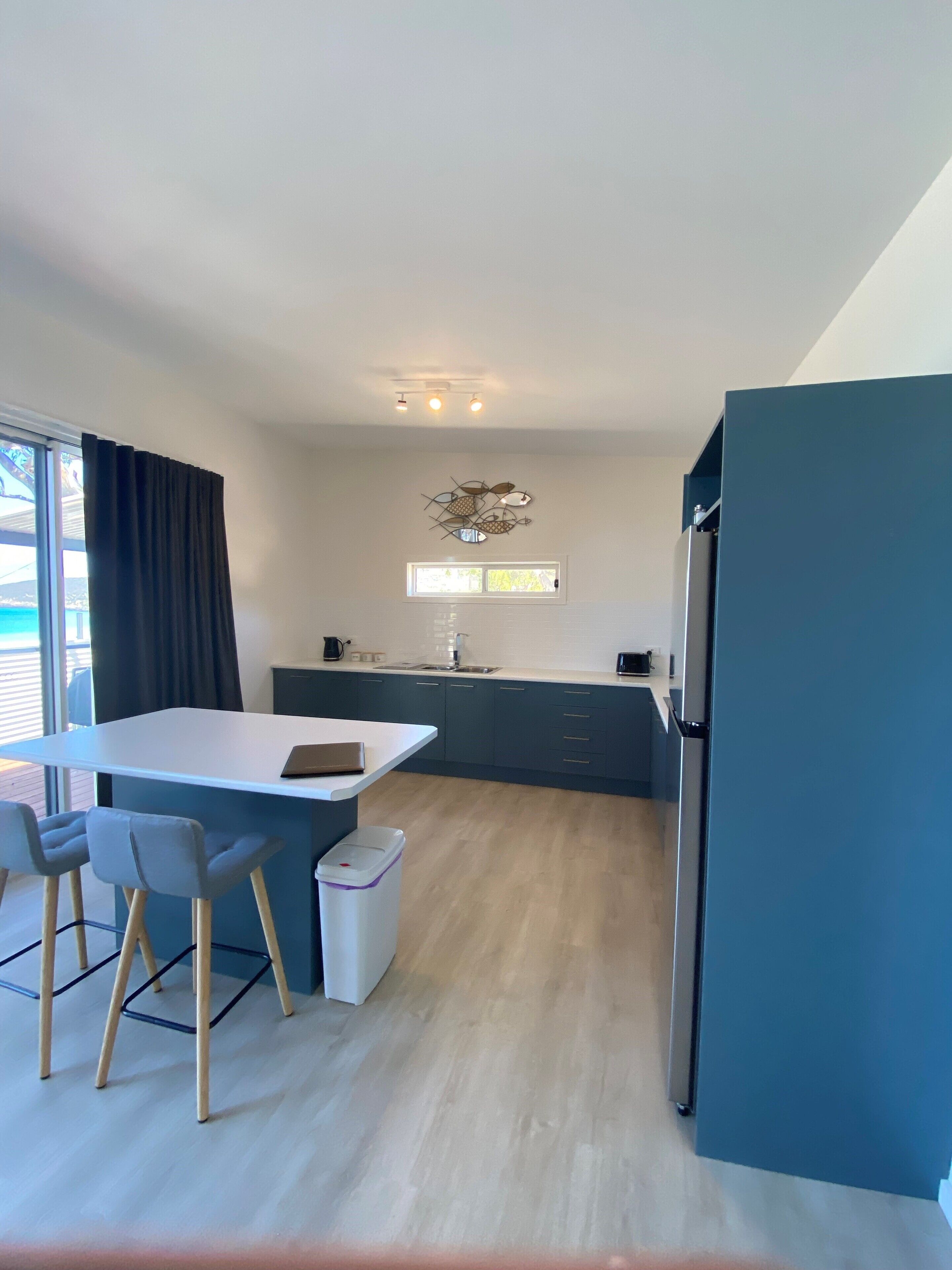 Una cocina moderna con encimera blanca, armarios azul oscuro y un refrigerador de acero inoxidable. Se observa una ventana con vistas al exterior y un pequeño contenedor de basura.