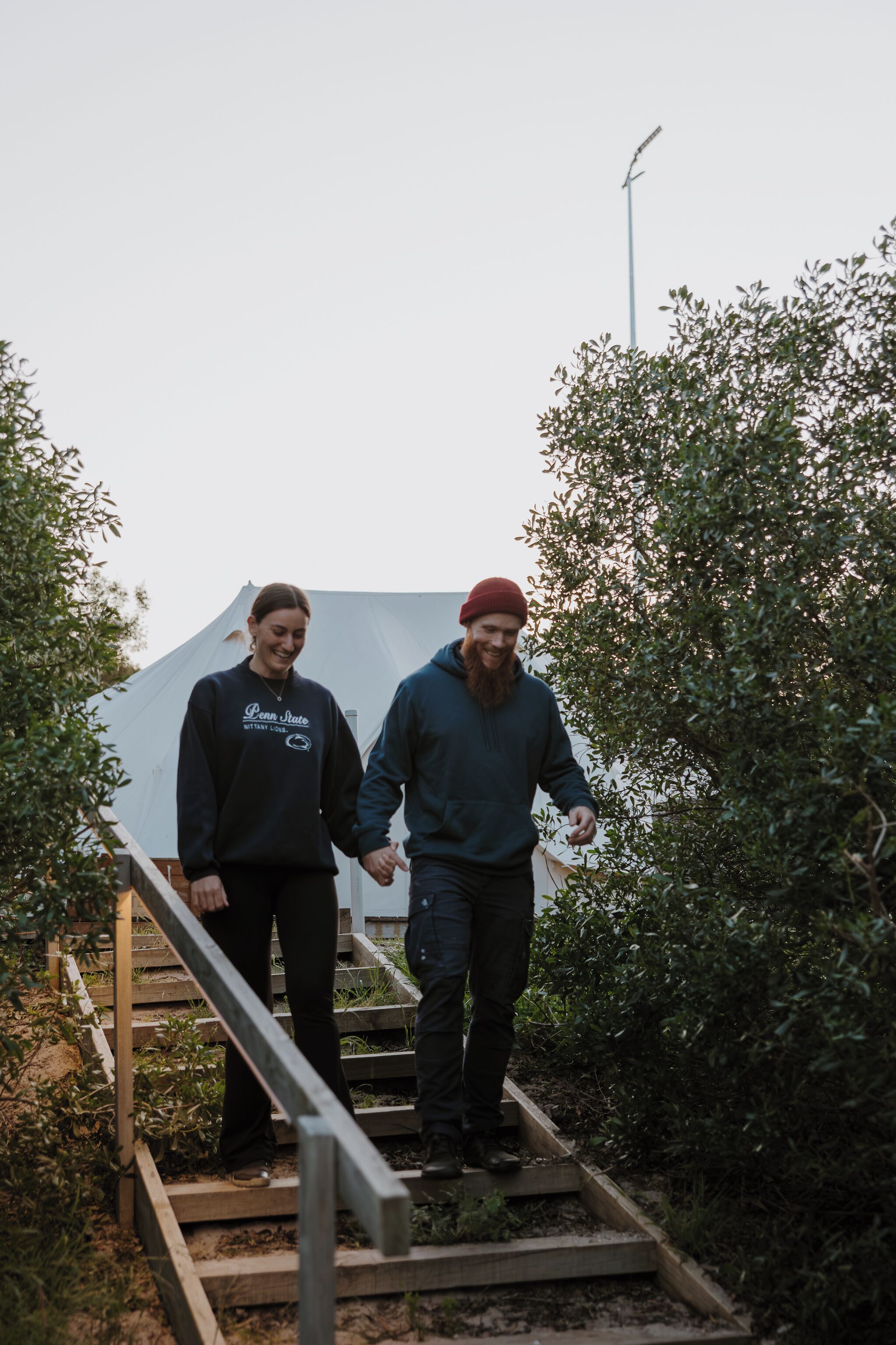 Twee personen lopen een houten trap op, omringd door groen.
