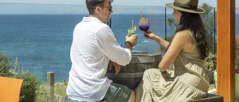 Two people seated at a table with a view of the ocean, on a deck with wicker furniture and a glass barrier.
