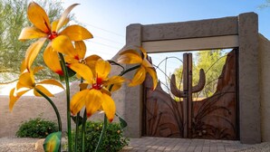 A metal gate with a cactus design, a paved entrance, and yellow flowers in the foreground. - Sprawling Arizona Oasis w/ Dream Backyard (New River)