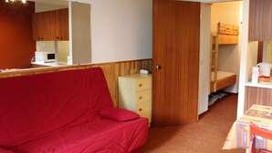 A compact room with a red sofa, a wooden wardrobe, a kitchenette, and a bunk bed.