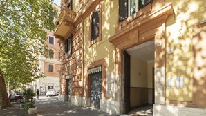A street view of a building with a large entrance, a number '5' sign, and a tree casting shadows on the pavement. - GuestHost - Comfortable and quiet 90sqm apartment that can accommodate up to 6 people. Located on the sixth floor, without lift (not suitable for the disabled, steps to access), of a renovated period building in Trastevere area, the apartment is located  (Roma)
