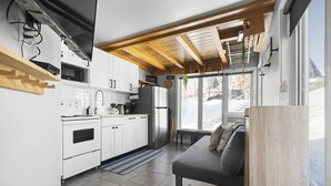A compact kitchen with white cabinets, a built-in oven, and a refrigerator. There is a gray sofa with cushions and a window providing a view of the outside. - Chouette Tremblant Ski In/Out Pedestrian Village (Mont-Tremblant)