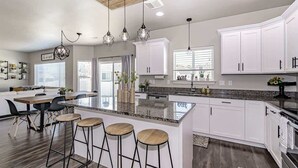 Una cocina moderna con armarios blancos, una isla central con taburetes, iluminación colgante y un ventanal grande.