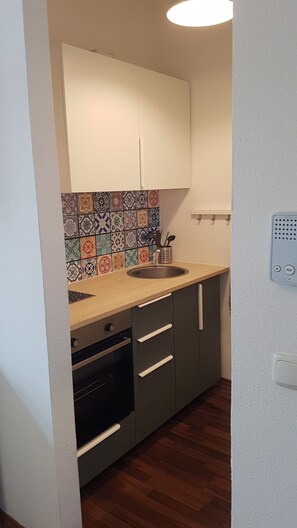 Una pequeña cocina con un lavamanos, estufa y microondas. Presenta un salpicadero de azulejos coloridos y una lámpara de techo blanca.