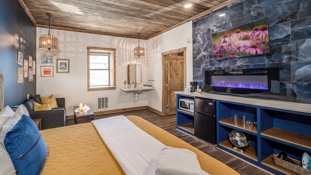 A modern hotel room with a bed, a fireplace, a television, and a kitchen area.