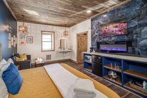 A modern hotel room with a bed, a fireplace, a television, and a kitchen area.