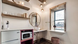 A compact kitchen with a refrigerator, microwave, and a sink. There are wooden shelves above the counter, and a window with a view of a bridge.