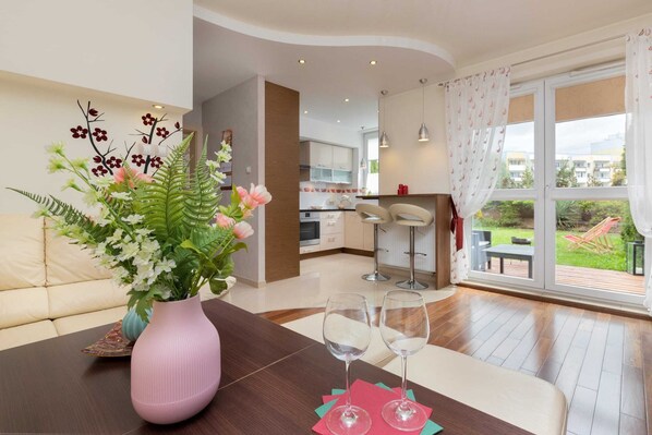 A modern living room with a sofa, a dining table with two wine glasses, a pink vase with flowers, and a view of the kitchen and outdoor area.