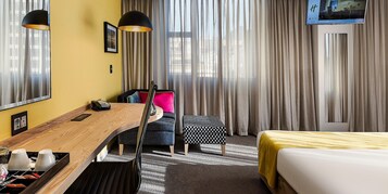 A hotel room with a wooden desk, a chair, a bed, a TV mounted on the wall, and a window with curtains.