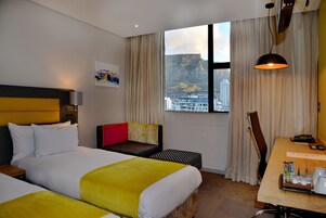 A hotel room with two beds, a desk, a chair, and a large window with a city view.