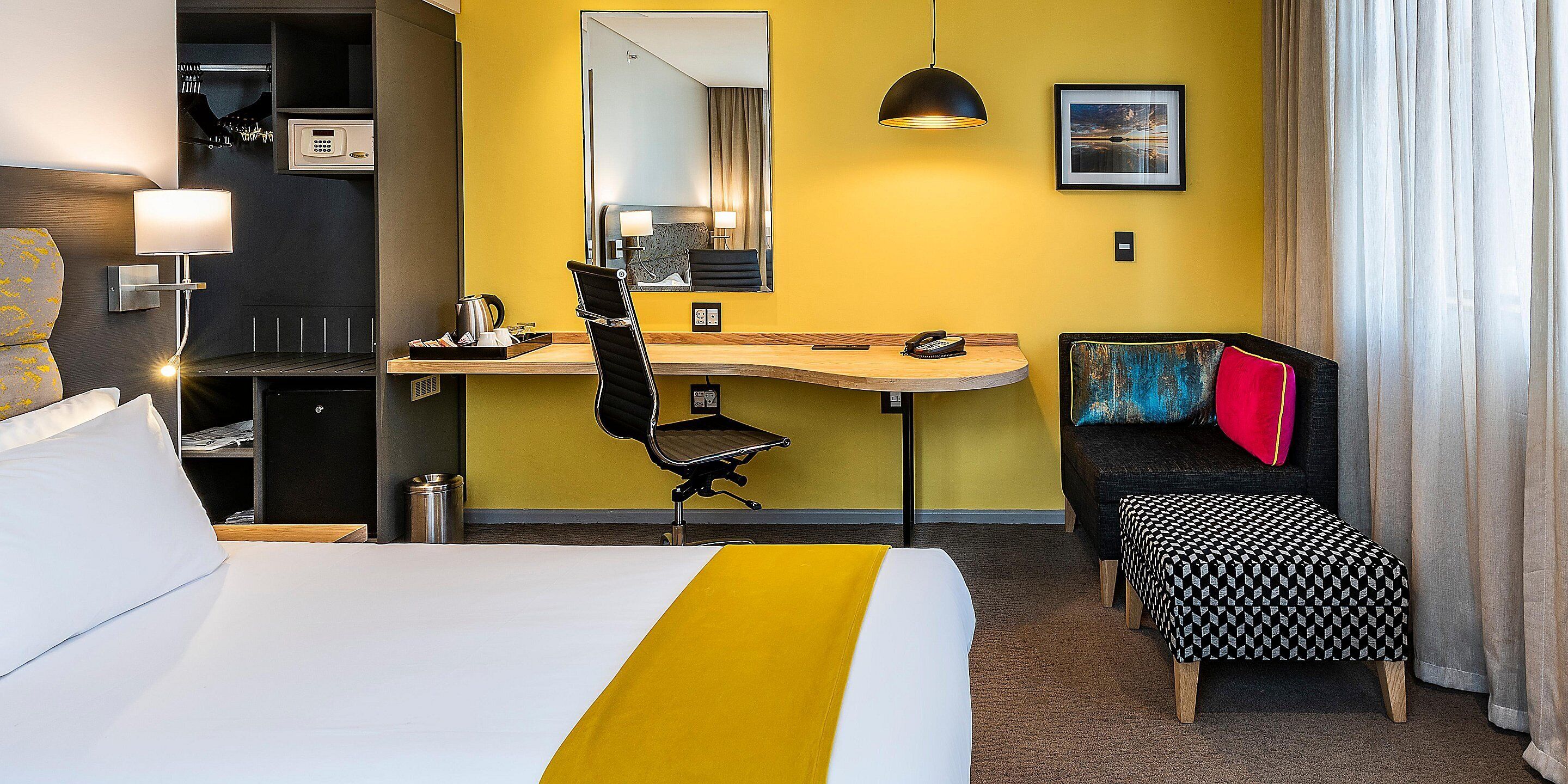 Ein Hotelzimmer mit Bett, Schreibtisch, Stuhl, kleinem Tisch, Bank, Spiegel, Lampe, einem Bild an der Wand und einem Fenster mit Vorhängen.
