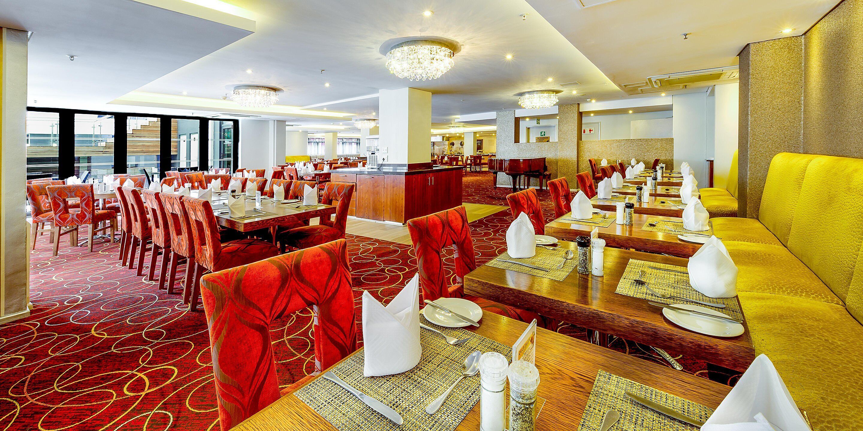 A hotel dining area with multiple tables and chairs, a piano, and a large chandelier.