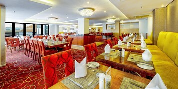 A hotel dining area with multiple tables and chairs, a piano, and a large chandelier.