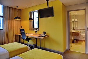 Une chambre d’hôtel avec deux lits, un bureau, une chaise, une télévision et une salle de bain visible à travers une porte ouverte.