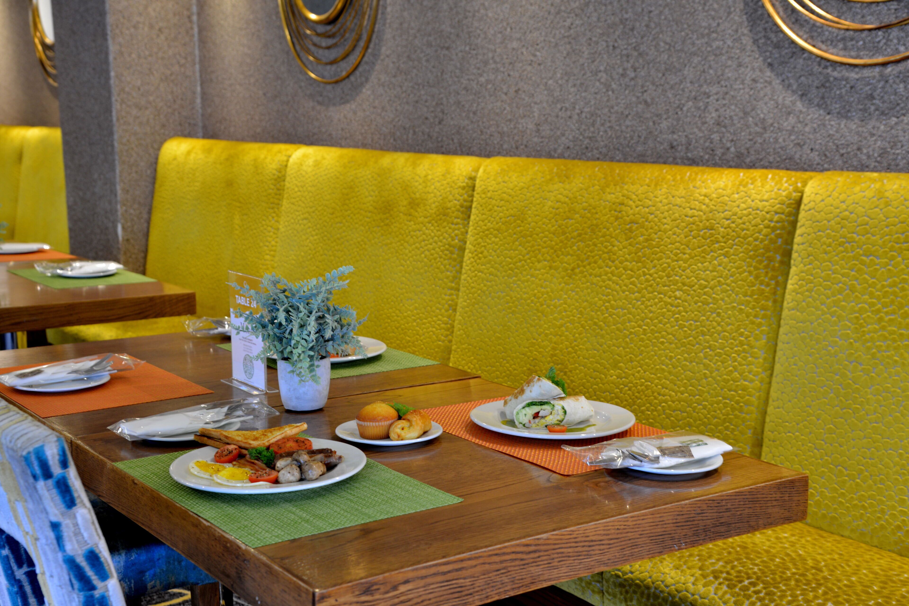 L’intérieur d’un restaurant avec des tables en bois, des banquettes jaunes et moelleuses, et différentes assiettes remplies de plats.