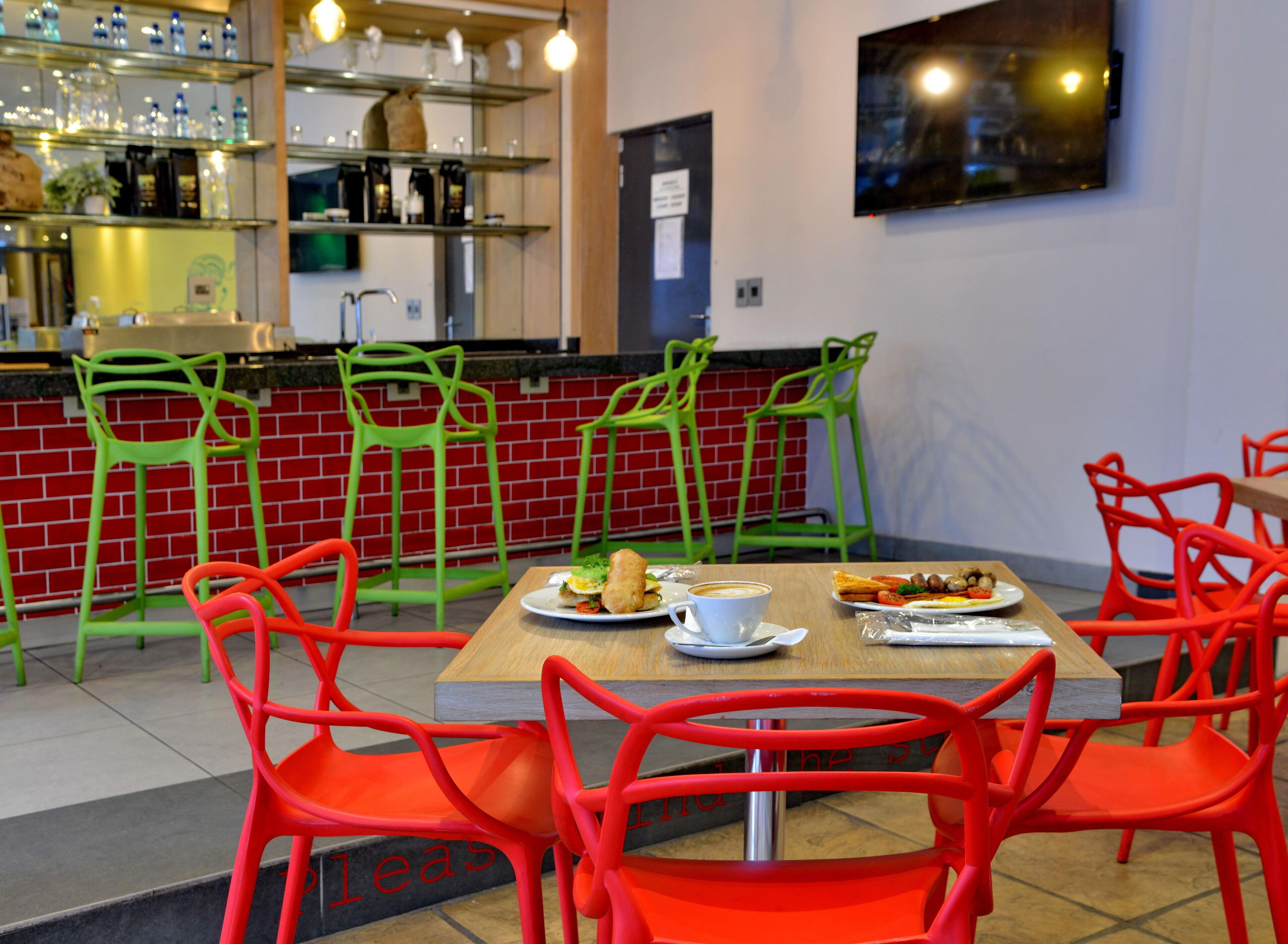 A restaurant with red and green chairs, a wooden table with plates of food, and a bar area with shelves of bottles and glasses.