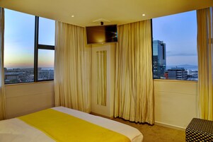 A hotel room with a large bed, a TV mounted on the wall, and a city view through large windows.