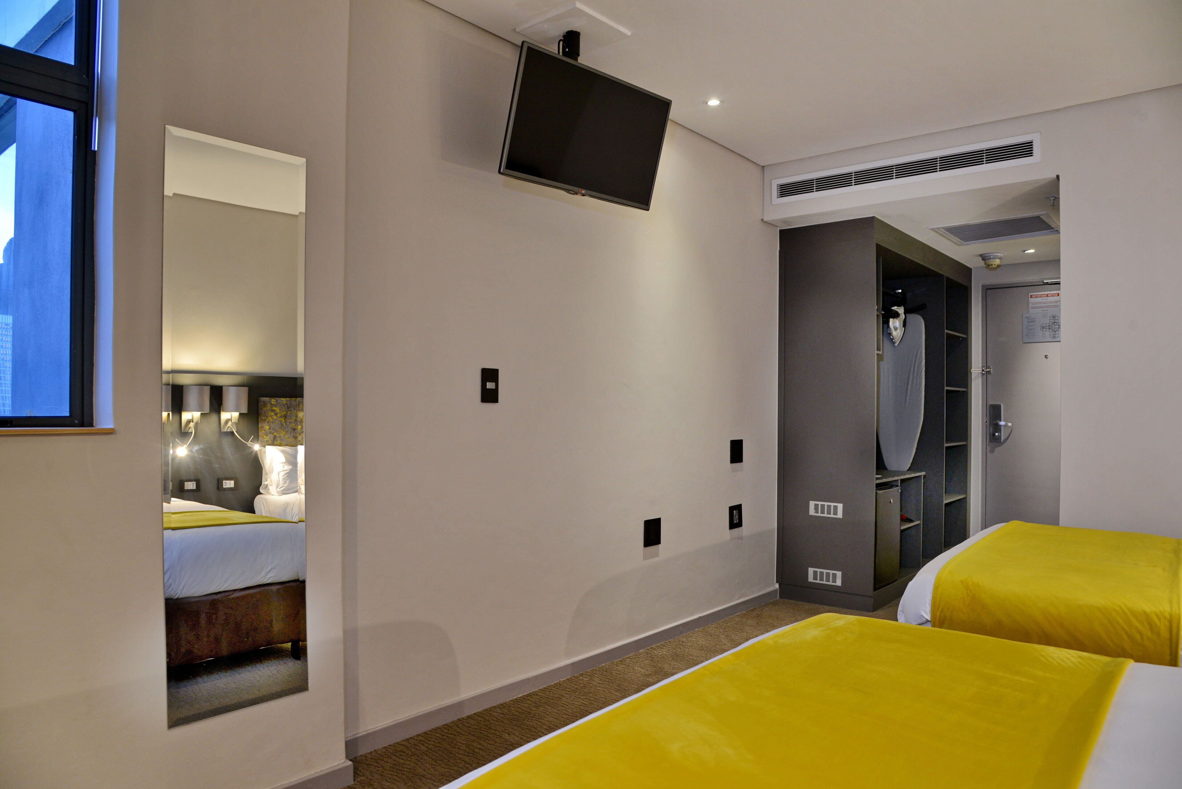 Quarto de hotel com duas camas, televisão presa na parede e um espelho refletindo o aposento.