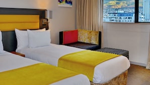 Une chambre d’hôtel avec deux lits, une grande fenêtre donnant sur la ville, un tableau accroché au mur et une fenêtre avec des rideaux.