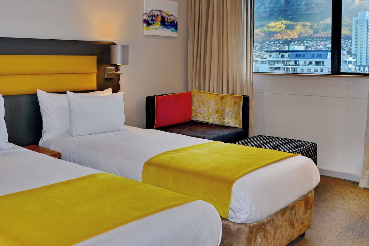 A hotel room with two beds, a large window with a city view, a painting on the wall, and a window with curtains.
