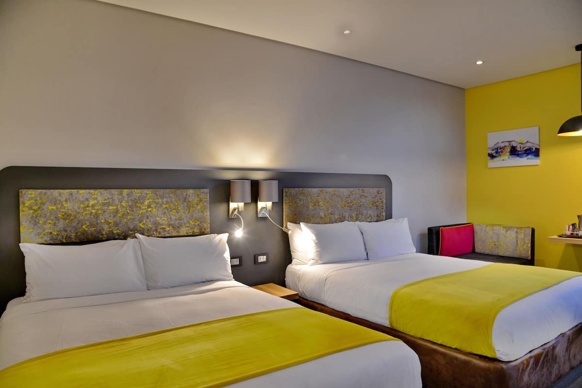 A hotel room with two beds, each with white linens and a yellow accent. There are wall-mounted lamps and a painting on the wall.