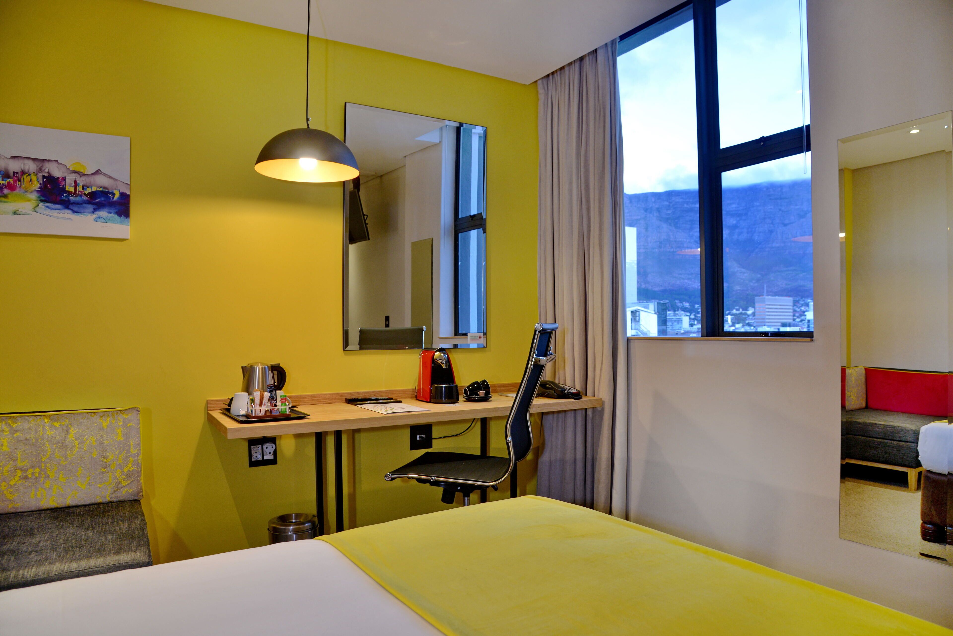 A hotel room with a bed, a desk with a chair, a coffee maker, a telephone, and a window with a city view.