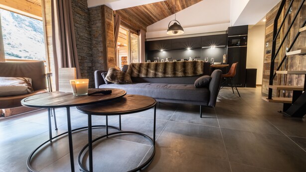 Een moderne woonkamer met een stenen muur, een grijze bank, een ronde salontafel, en een eethoek met een houten tafel en stoelen.