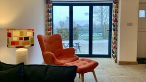 Un salón con un ventanal grande, un sillón moderno color naranja, una lámpara colorida y una pared de piedra.