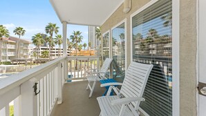 A balcony with white chairs and a pool view, surrounded by palm trees and buildings.