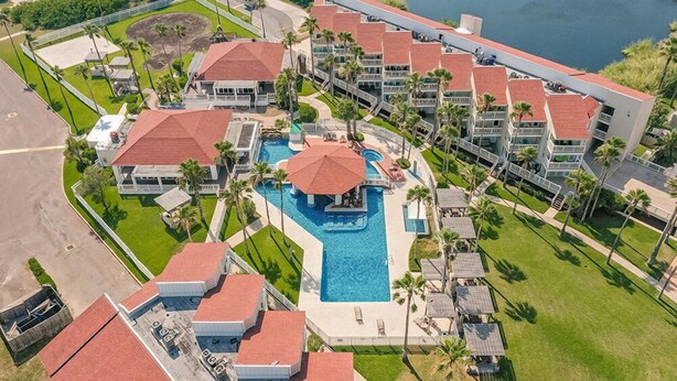 An aerial view of a resort with multiple buildings, a central swimming pool with a red roof, and surrounding palm trees.