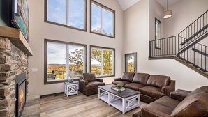 A spacious living room with a stone fireplace, large windows, a brown leather sofa, and a staircase. - NEW! Luxury Lakefront Private Lodge • Resort Pools • Workout Gym • Over 30 FREE amenities! (Kimberling City)