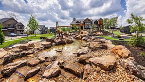 A residential area with a small stream, wooden play structures, and houses with large windows. - NEW! Luxury Lakefront Private Lodge • Resort Pools • Workout Gym • Over 30 FREE amenities! (Kimberling City)