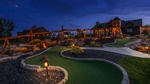 A well-lit golf course at dusk with a wooden bridge, rocks, and a house in the background.