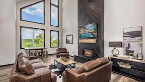 A spacious living room with leather sofas, a fireplace, a television, and large windows offering a view of greenery.