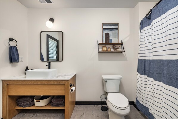 Un baño con un lavamanos blanco, tocador de madera, inodoro y cortina de ducha.