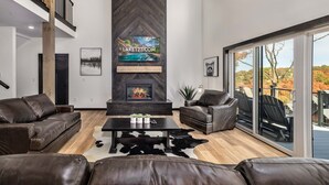 A modern living room with leather sofas, a fireplace, and a large window overlooking a deck. - Luxury Lakefront Lodge | 6BR Modern Retreat + FREE Tickets, Pools & Amenities (Kimberling City)