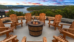 Un foyer circulaire en bois, entouré de chaises Adirondack, offre une vue sur un lac et des collines boisées.