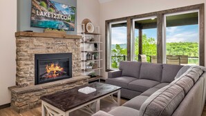A living room with a stone fireplace, a gray sofa, and a large window overlooking a green landscape. - Family-Friendly Lake Cabin | Hot Tub + King En-Suites + Lake Access (Kimberling City)