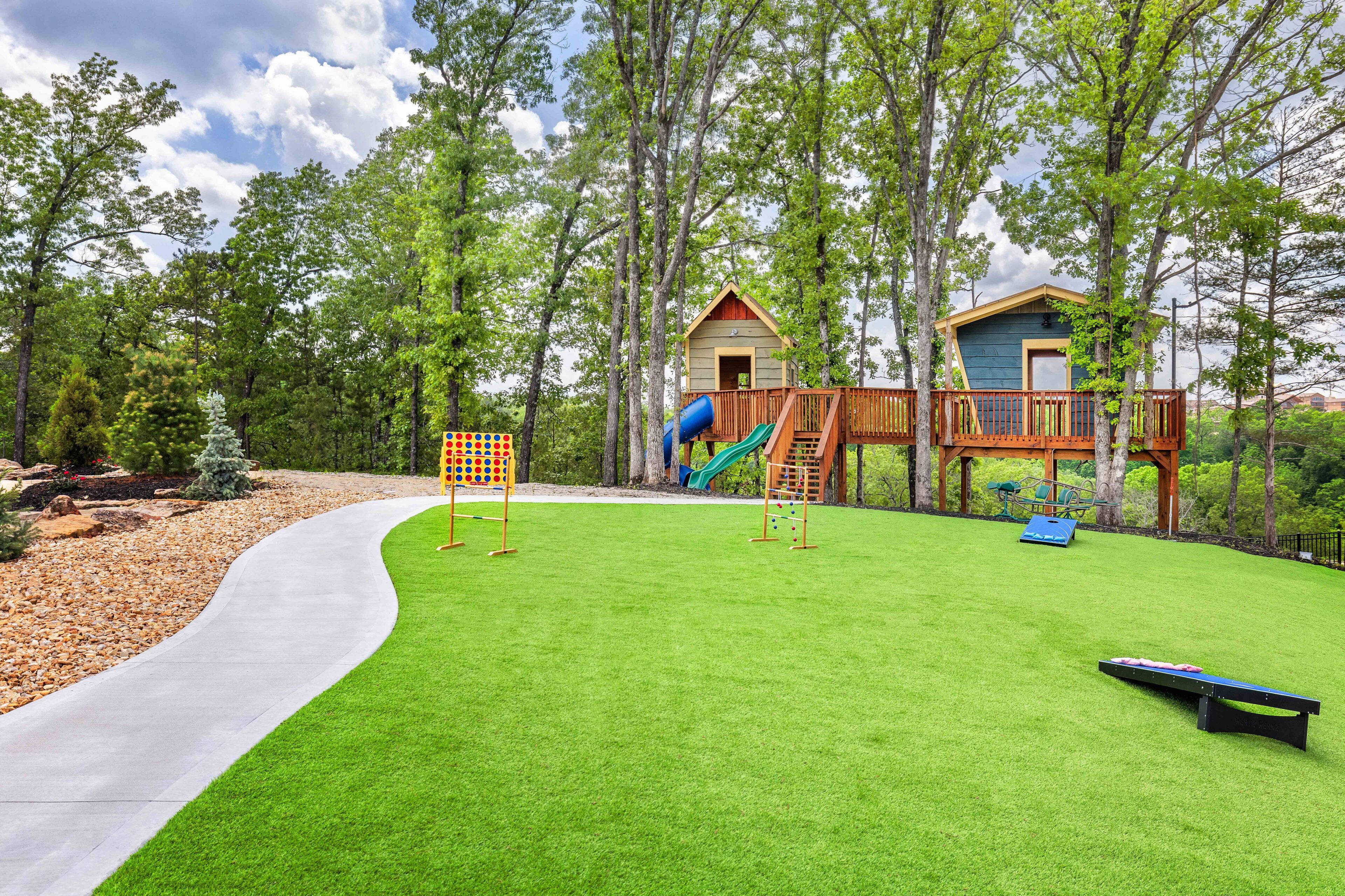 Ein Spielplatz mit Rutsche, Schaukeln und einer Kletterkonstruktion, umgeben von Bäumen und einem befestigten Weg.