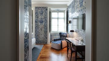 A narrow hallway with blue patterned tiles, a wooden floor, a window with blinds, a small seating area with a chair and ottoman, and a table with a lamp and decorative items.