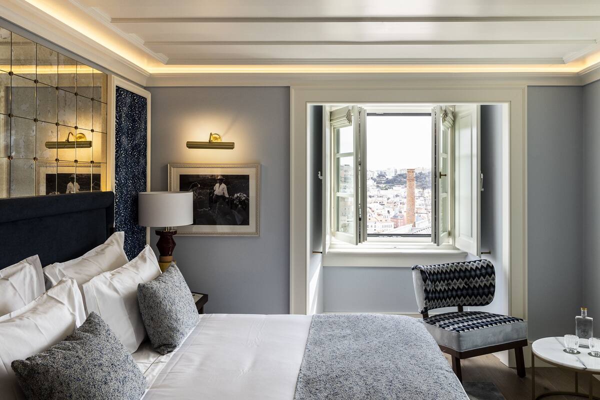 Ett hotellrum med en säng, en stol, ett litet bord, ett fönster med utsikt, en öppen spis och en spegel som hänger på väggen.