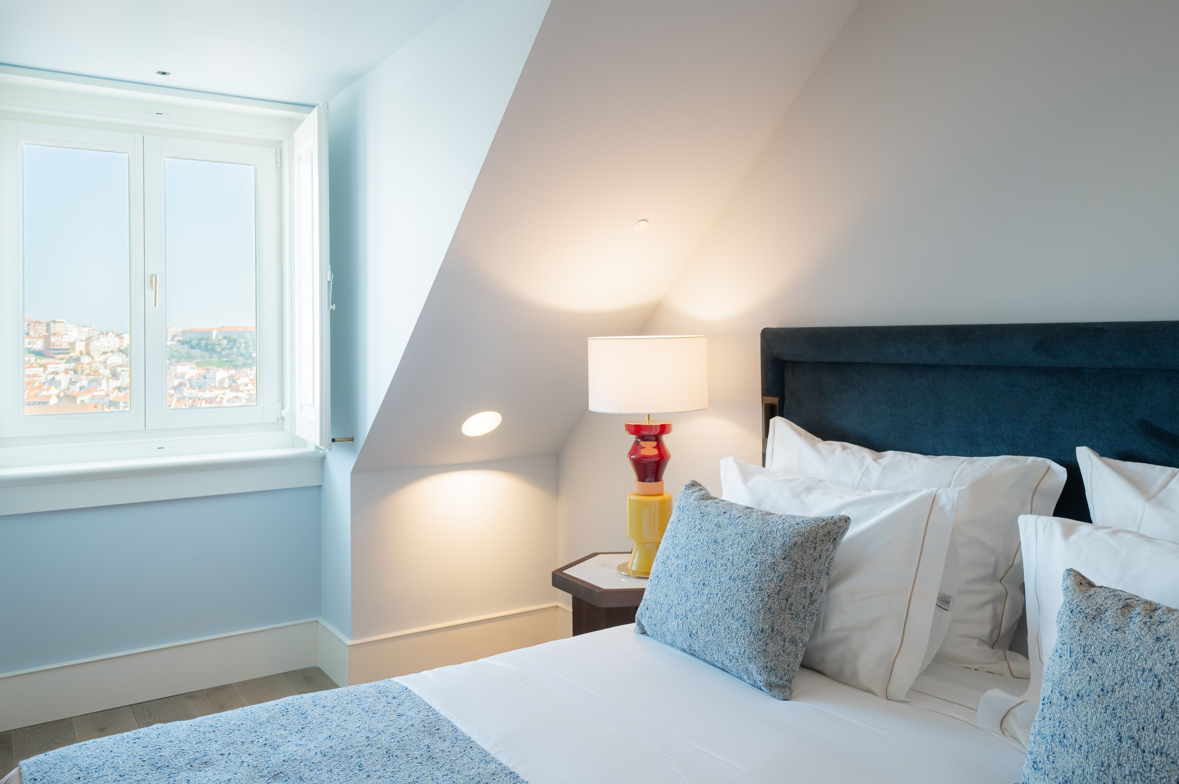 A bedroom with a bed, pillows, a nightstand, a lamp, and a window with a view.